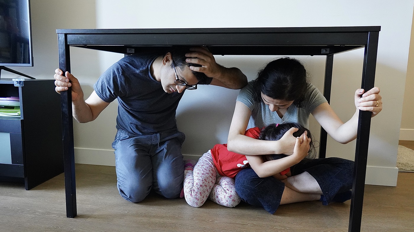 A man, woman, and child take cover under a table during an earthquake. The man and woman are holding onto the table with one hand.