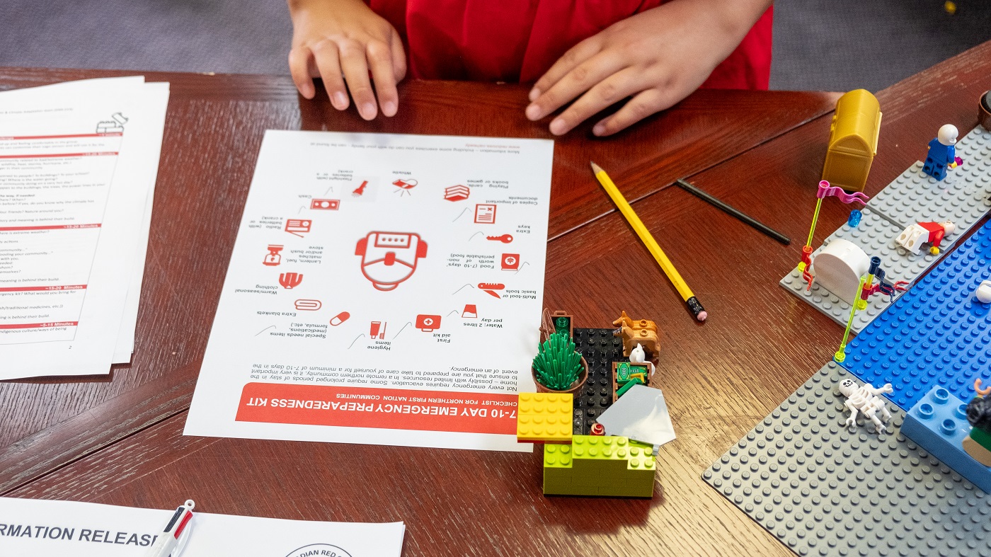 Les mains d'un enfant sont appuyées sur une table devant une feuille de papier présentant des icônes d'articles à mettre dans une trousse d'urgence. On voit des blocs LEGO sur le bord de la table.